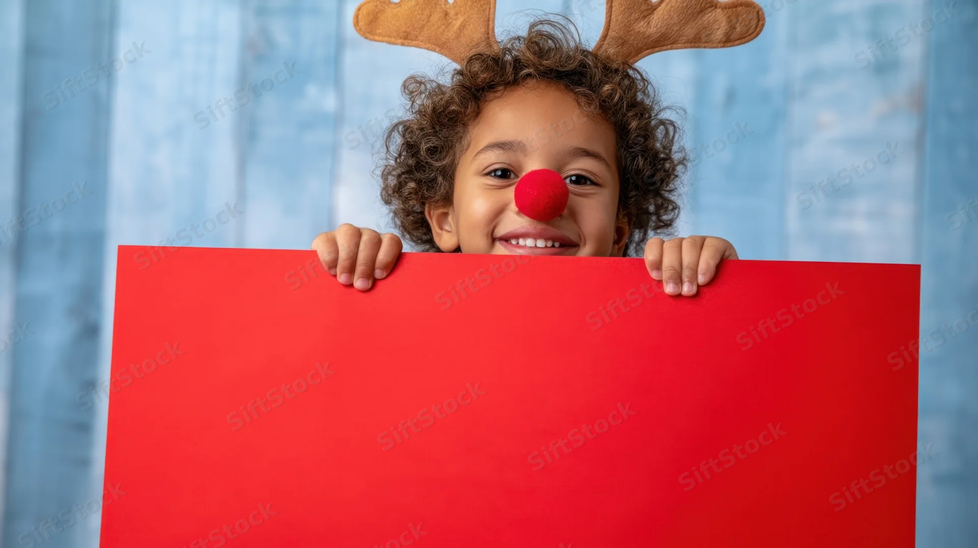 Young child peeking from behind red poster board with reindeer antlers and red nose, smiling, blue wooden background