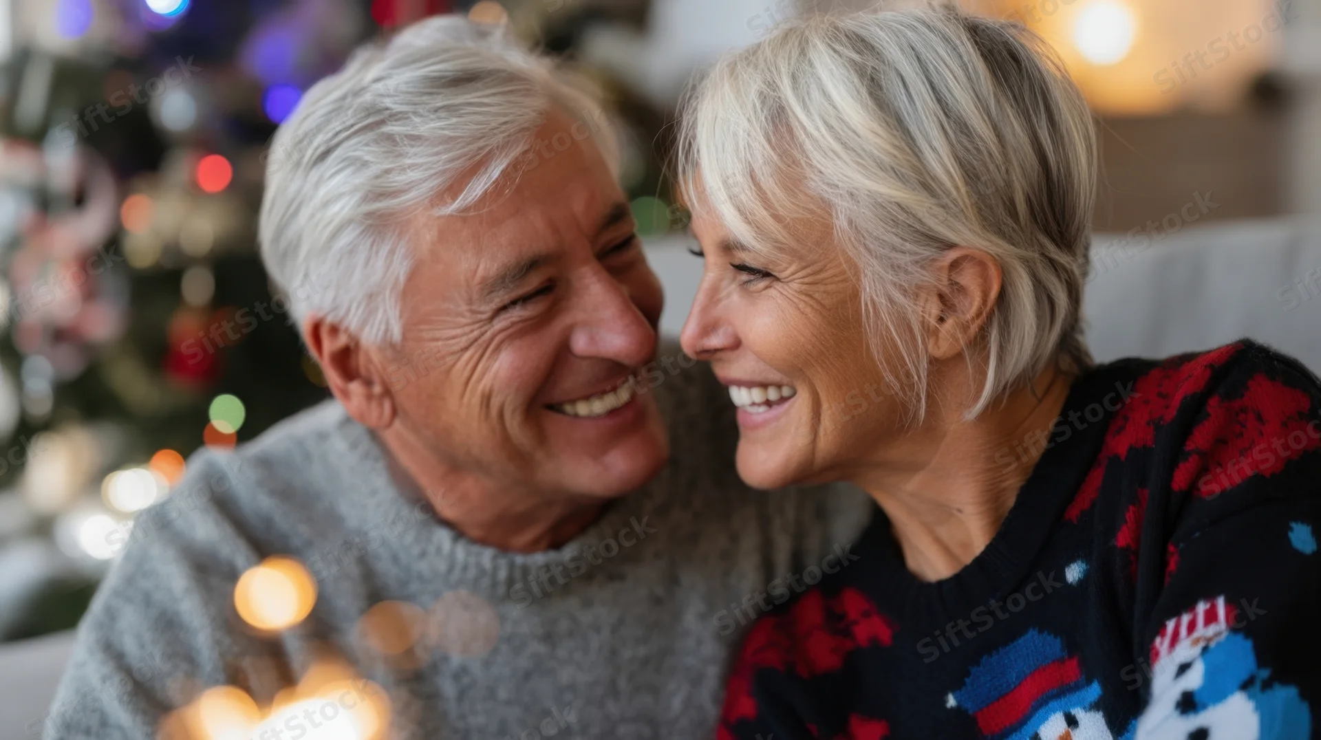 Elderly couple smiling warmly at each other in a cozy living room with christmas lights in the background
