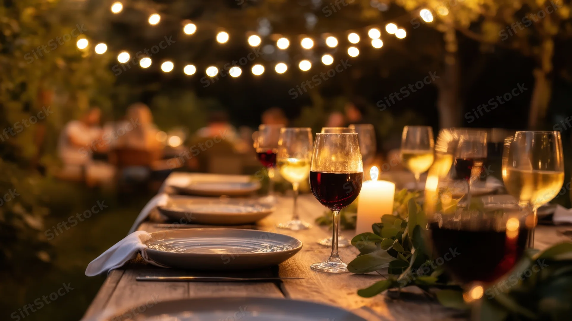 Long wooden table set with wine glasses and candles under string lights in a garden at evening