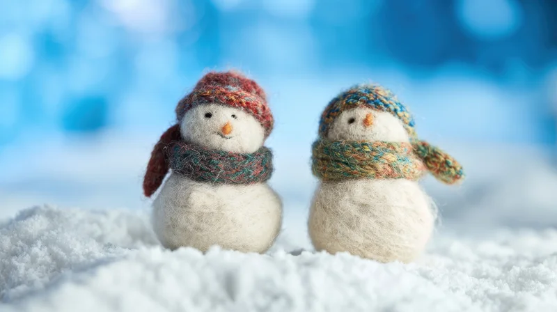 Two small felt snowmen wearing knitted hats and scarves standing in snow against a blue blurred background