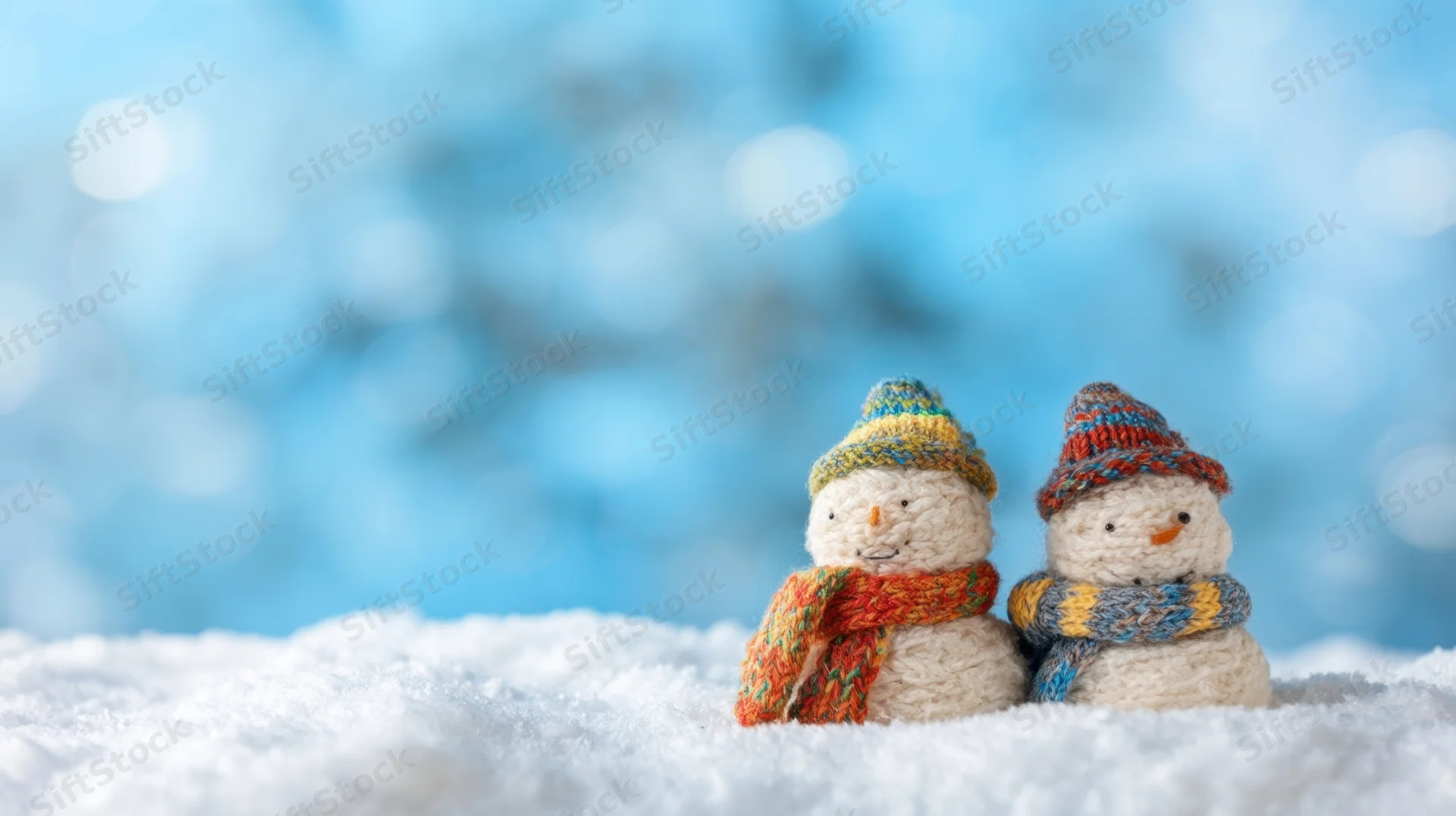 Two knitted snowmen wearing colorful hats and scarves sitting on snow against a blue bokeh background