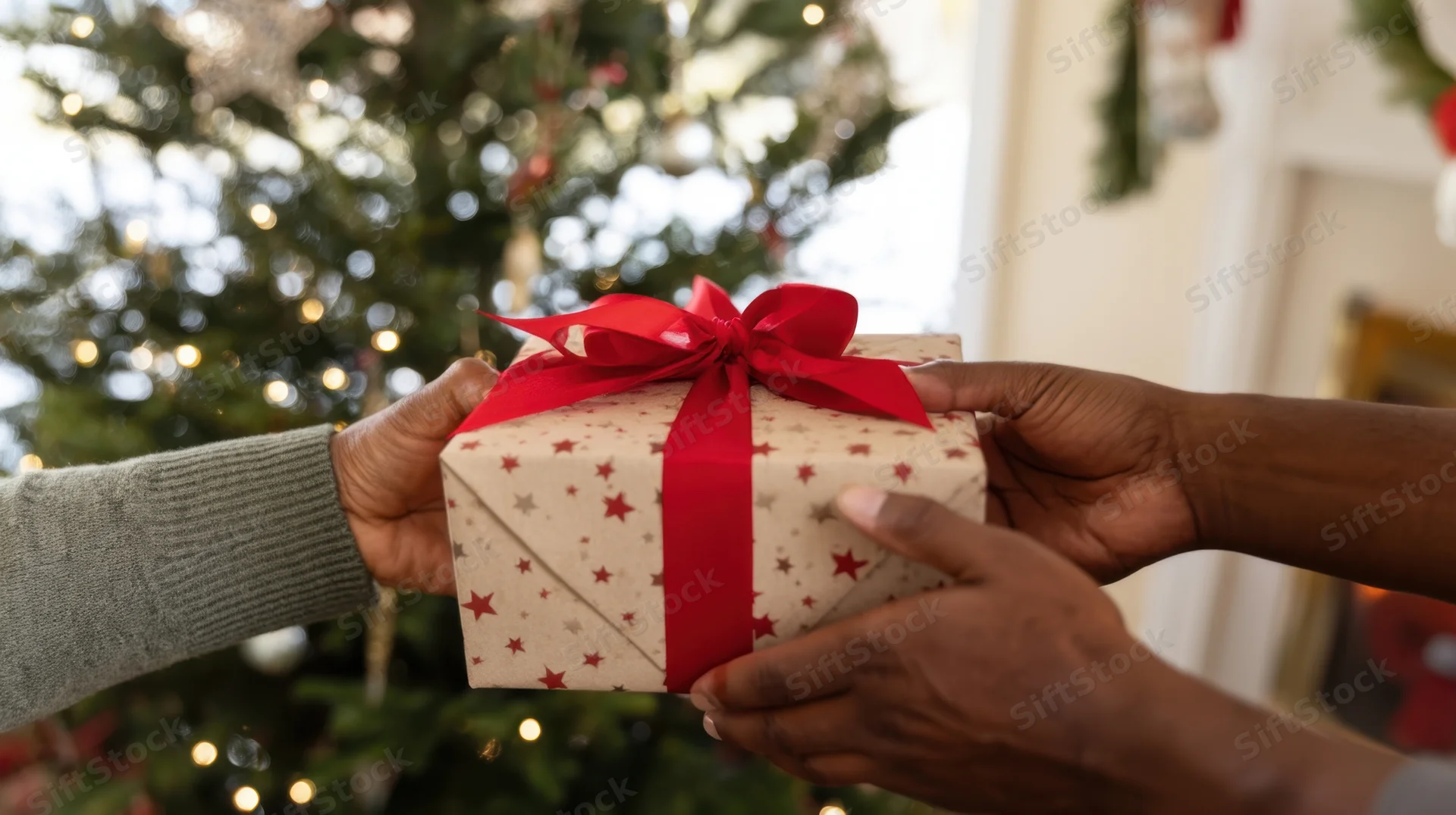 Hands exchanging a wrapped gift with red ribbon and stars in front of a christmas tree