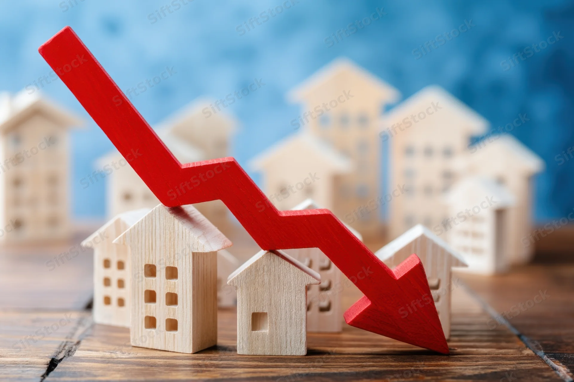 Red downward arrow resting on miniature wooden houses on a wooden surface