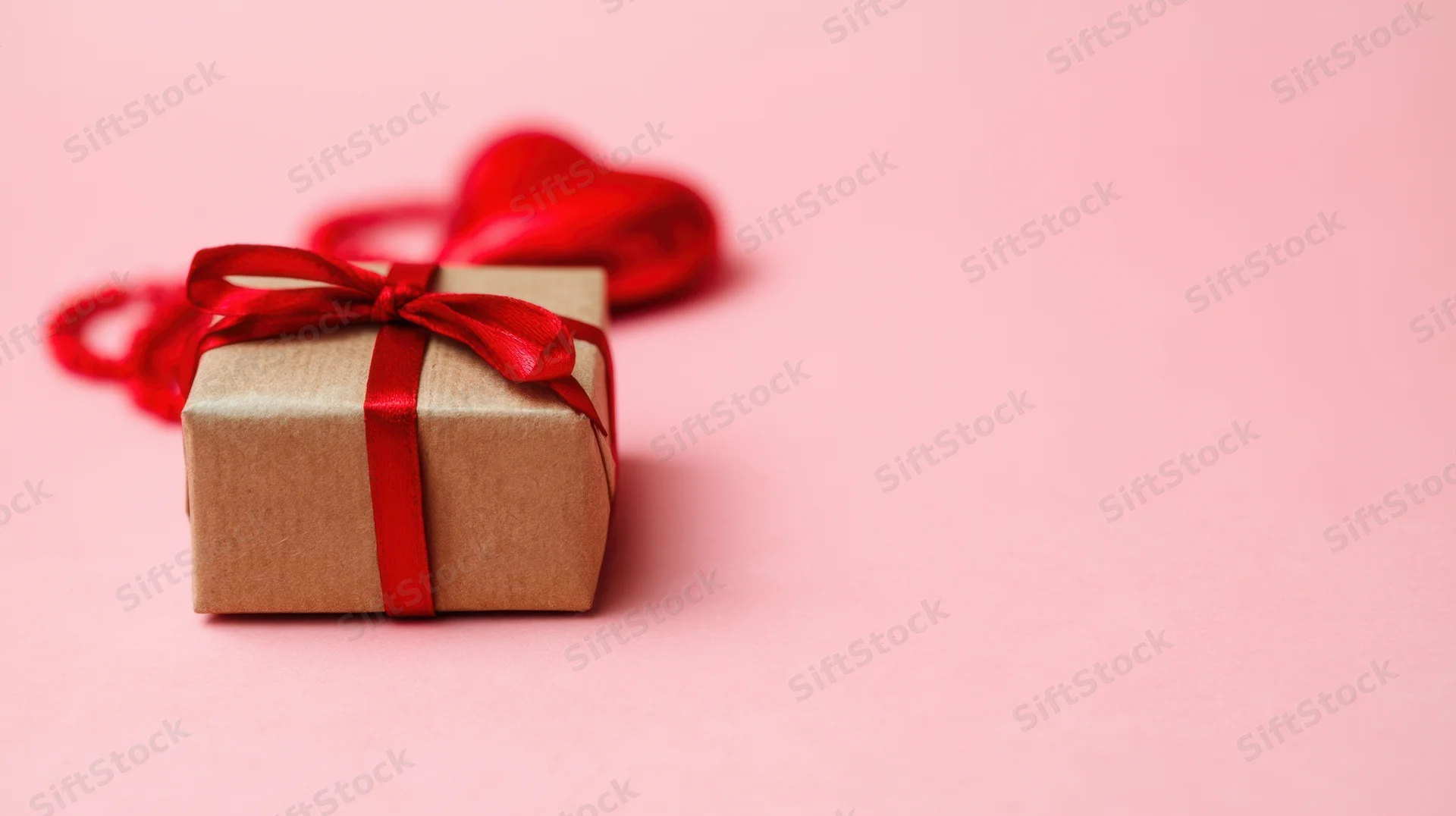 Gift box wrapped in brown paper with red ribbon and heart decoration on pink background
