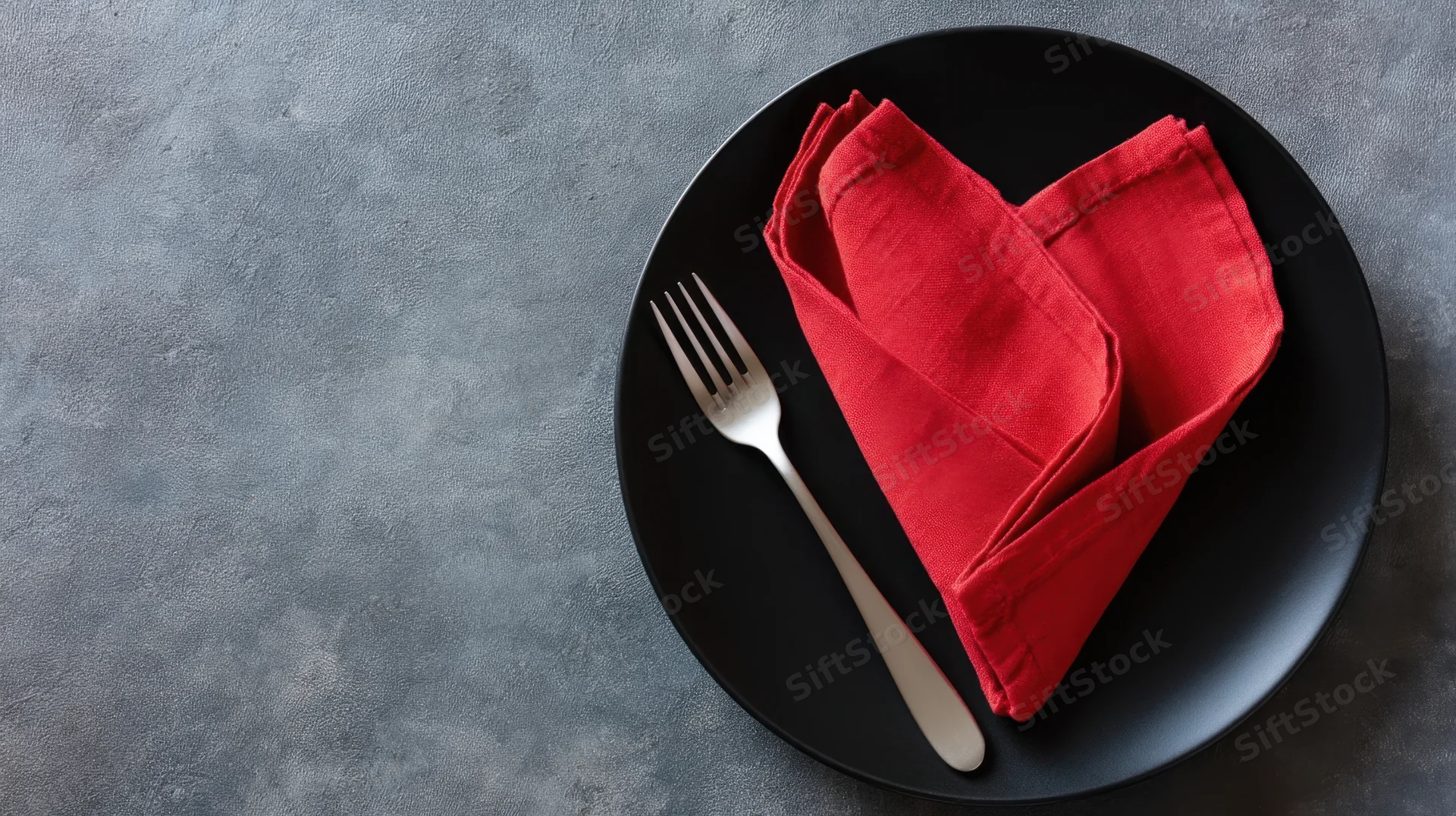 Red napkin folded into heart shape on black plate with fork, set on gray textured surface