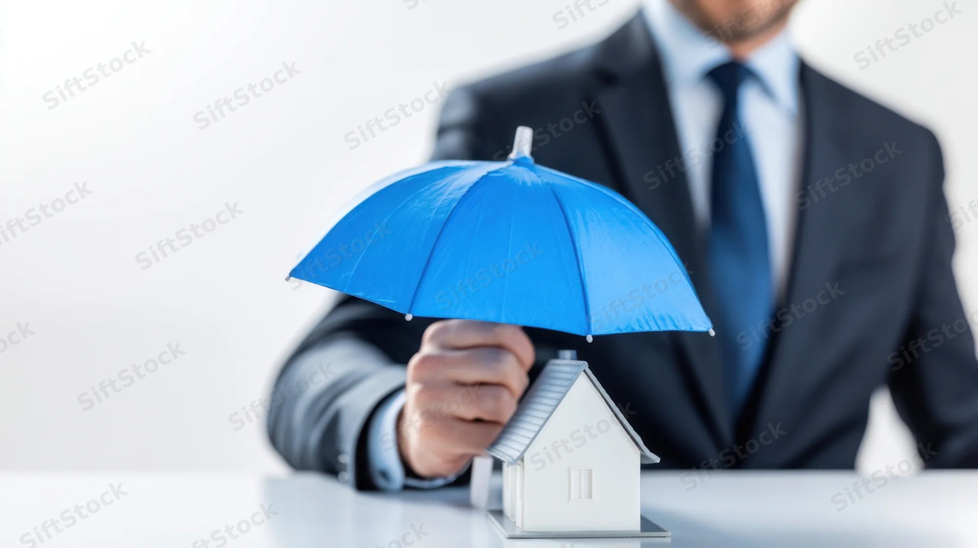 Businessman holding blue umbrella over miniature house on desk