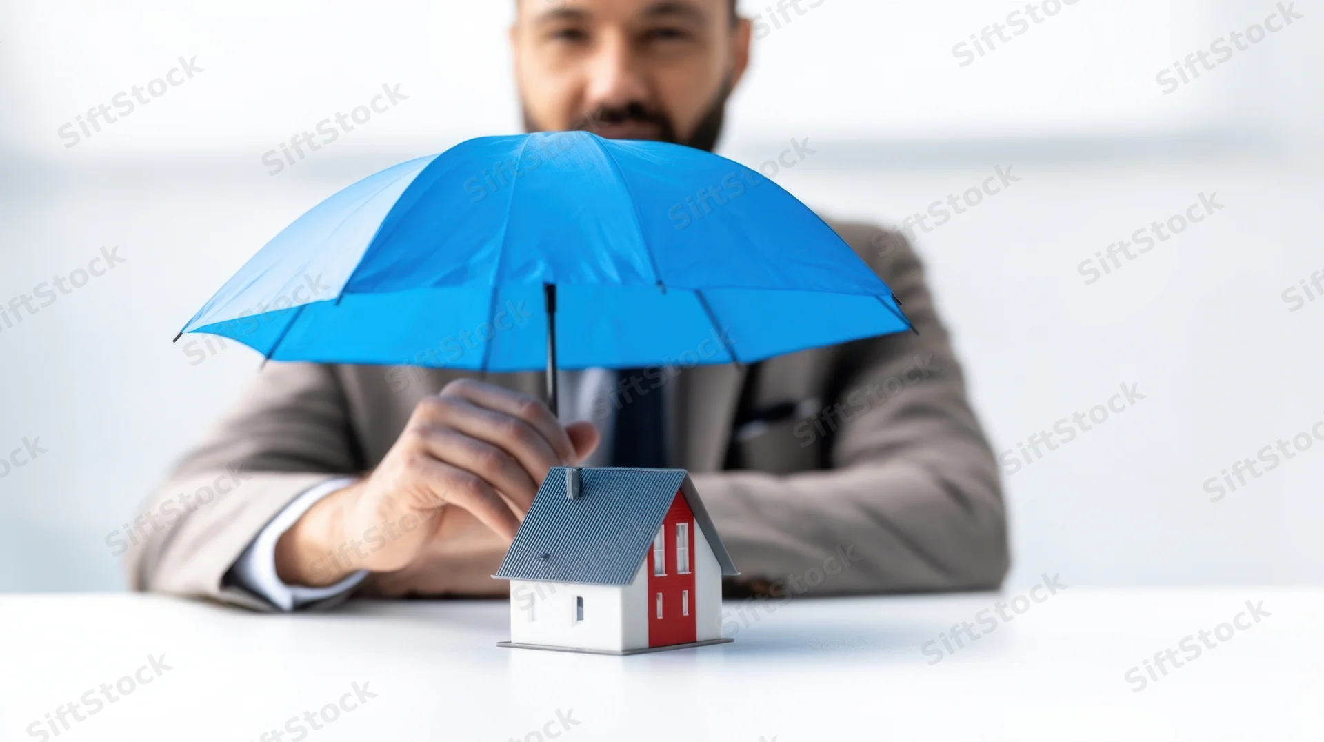 Man in suit holding blue umbrella over small house model on desk