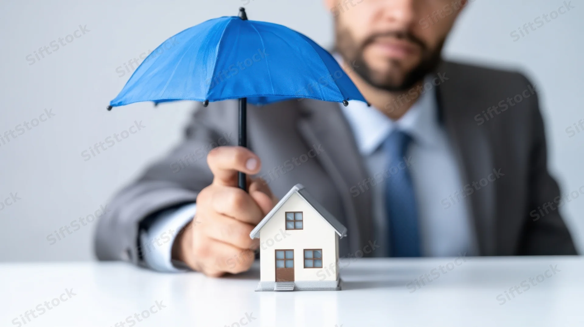 Man in suit holding blue umbrella over small house model on desk