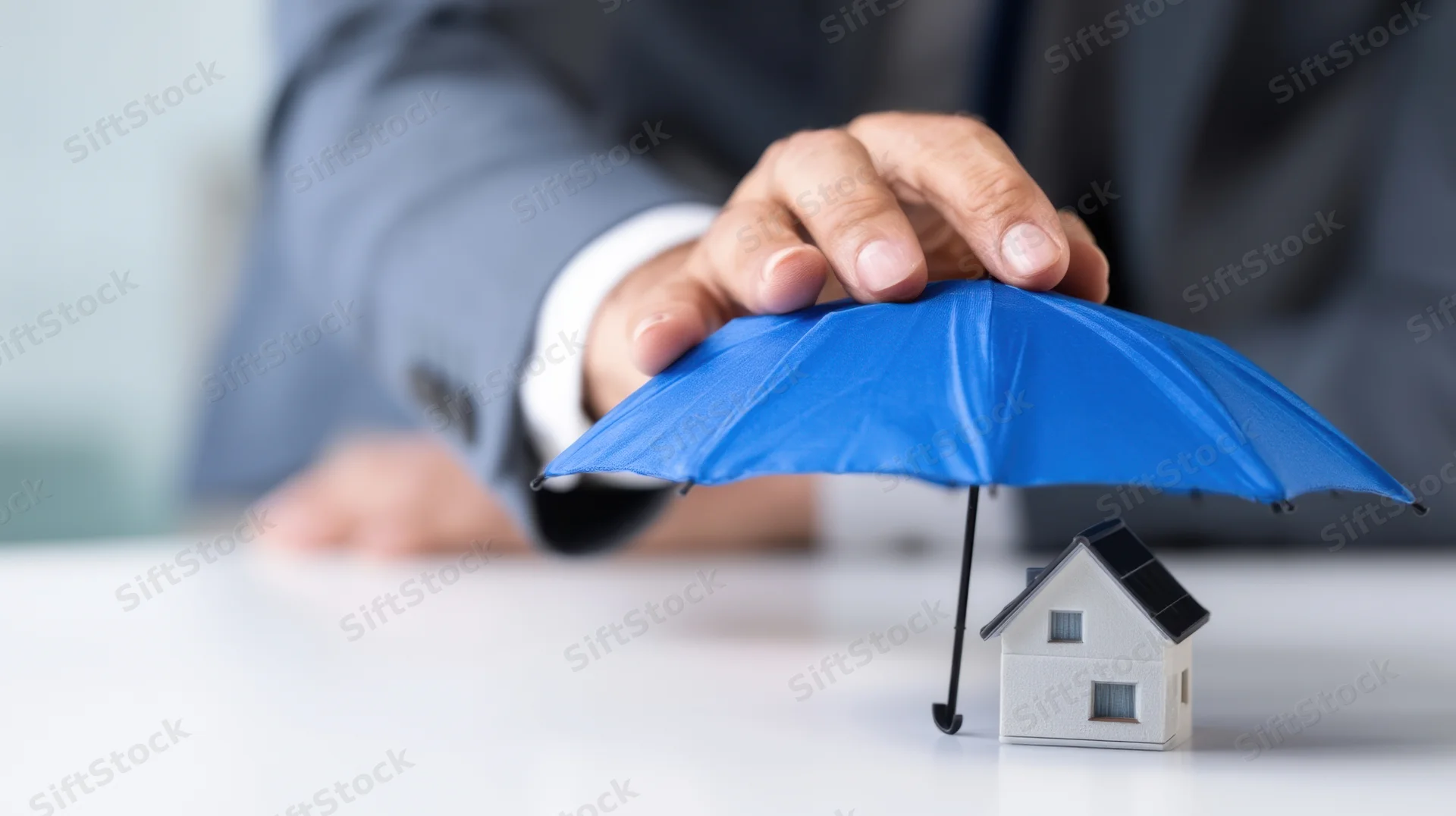 Businessman's hand holding blue umbrella over miniature house on desk