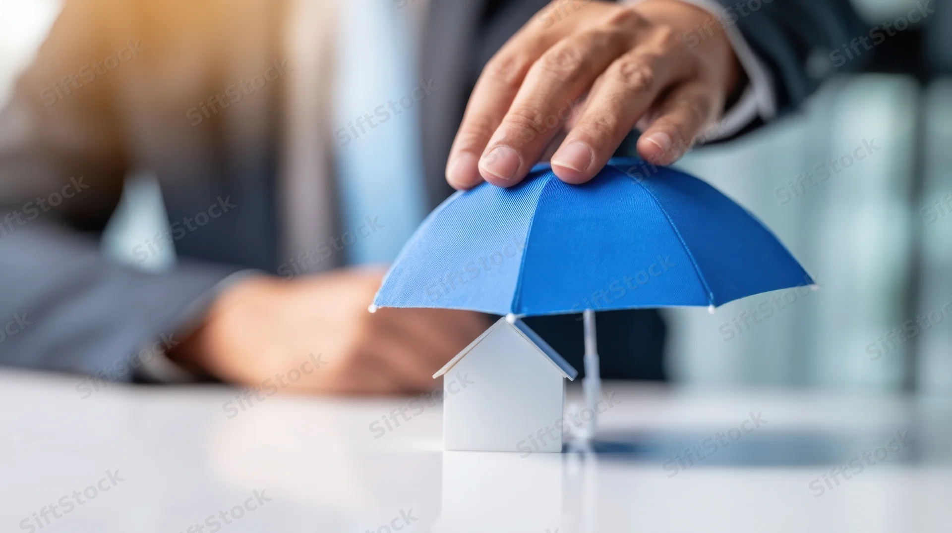 Hand placing blue umbrella over white house model on desk