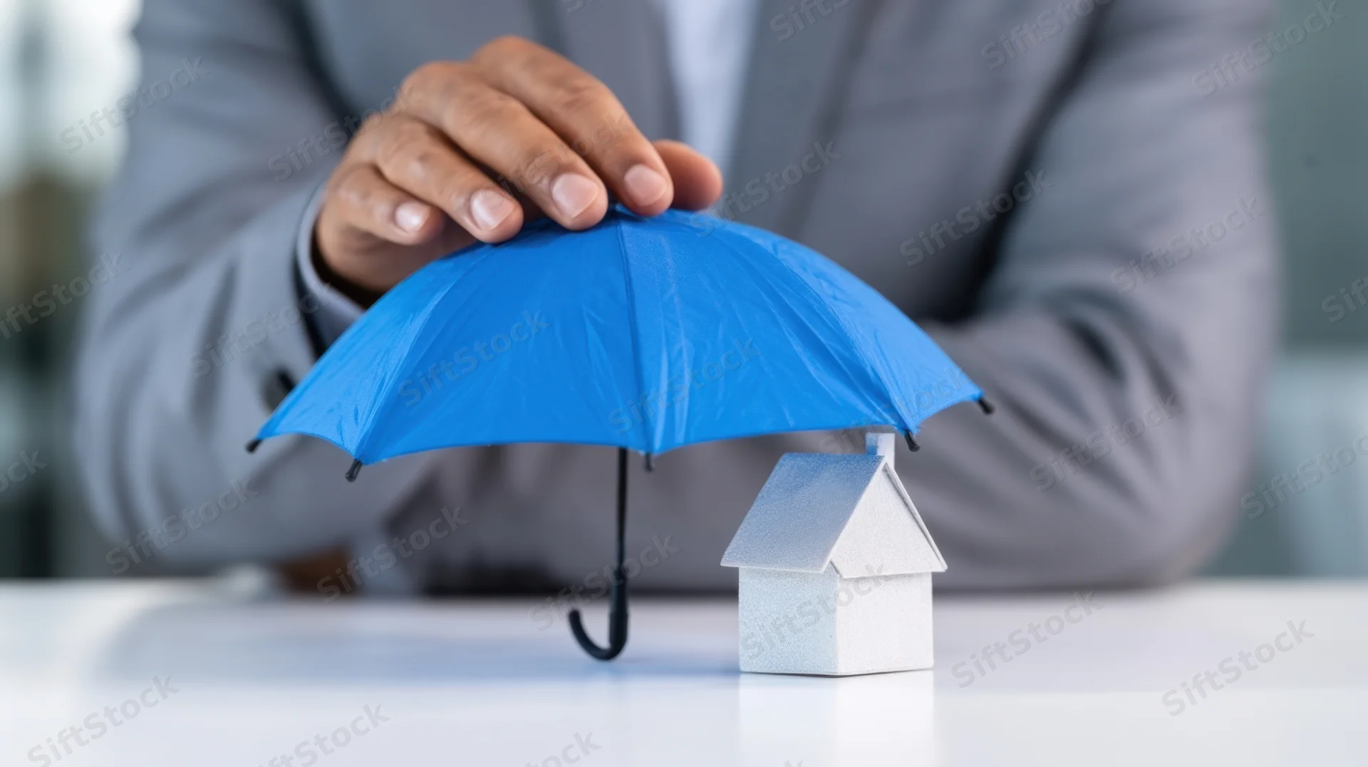 Businessman holding blue umbrella over miniature house on desk