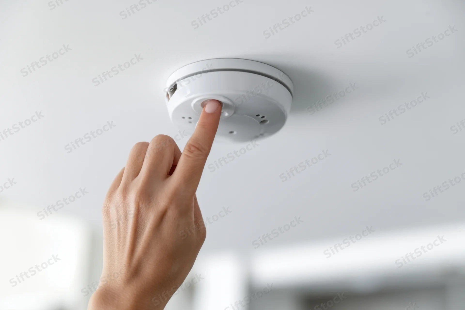 Hand pressing button on white smoke alarm mounted on ceiling
