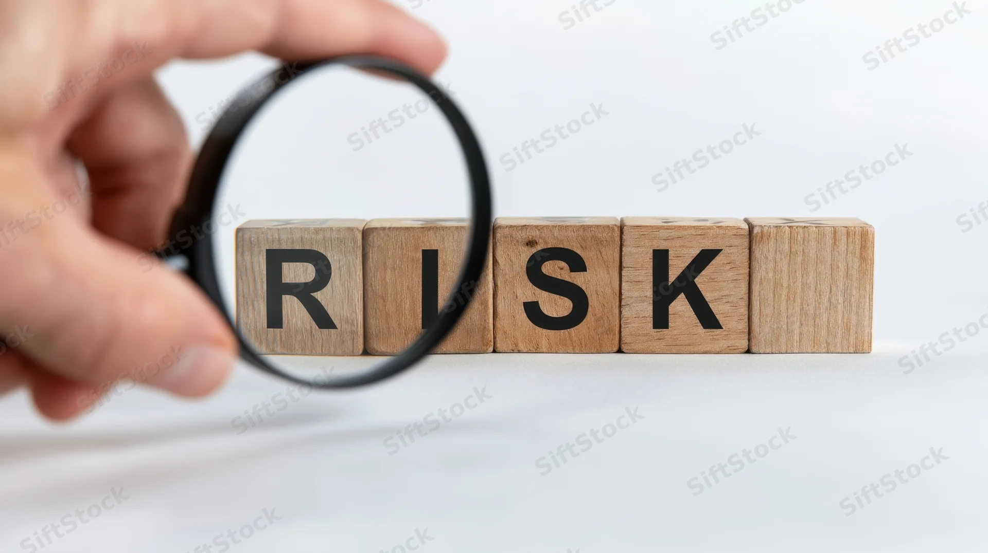 Hand holding magnifying glass over wooden blocks spelling risk on white surface