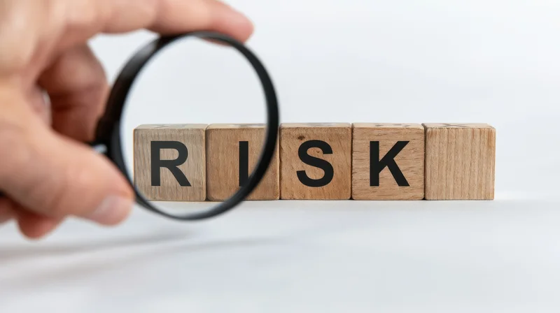 Hand holding magnifying glass over wooden blocks spelling risk on white surface