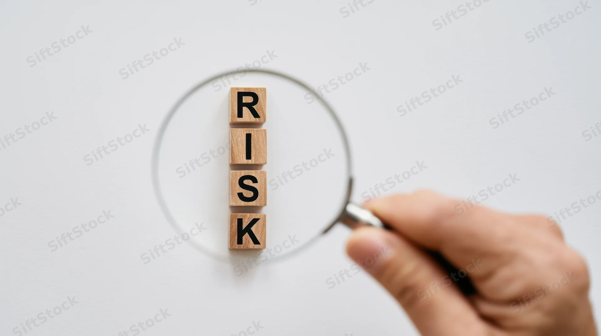 Hand holding magnifying glass over wooden blocks spelling risk on white surface