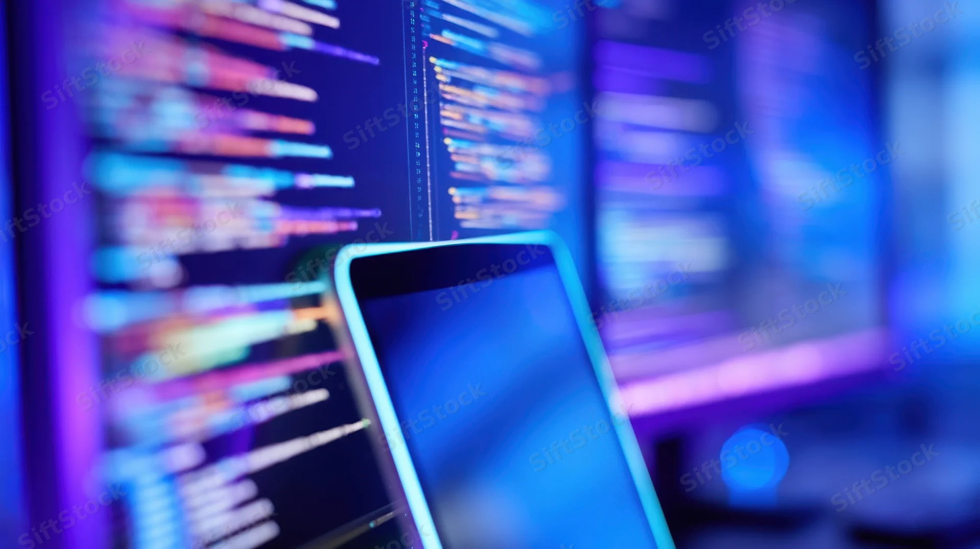Smartphone screen in focus with blurred code on computer monitors behind it