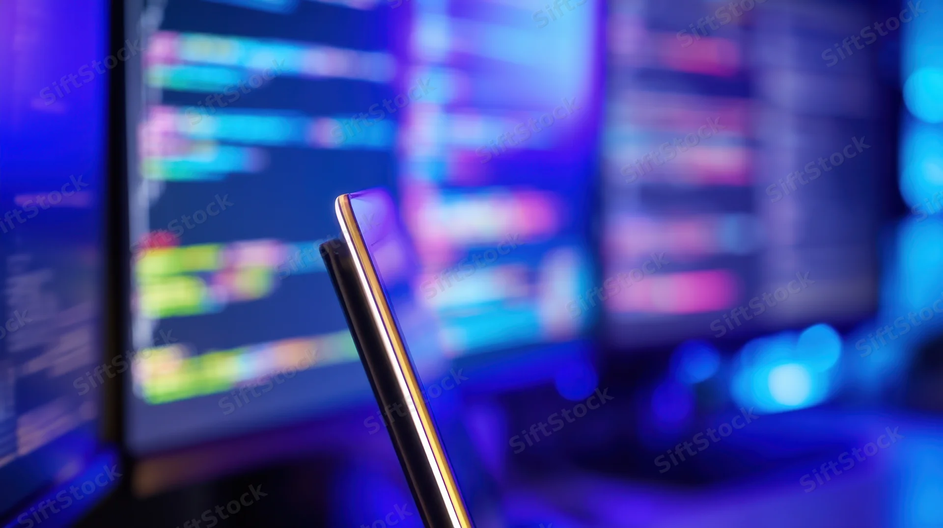 Smartphone screen angled in front of blurred computer monitors displaying colorful code in a dark room