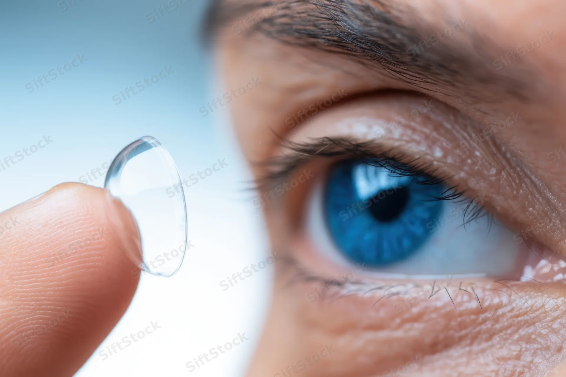 Close-up of a blue human eye next to a contact lens held between fingers