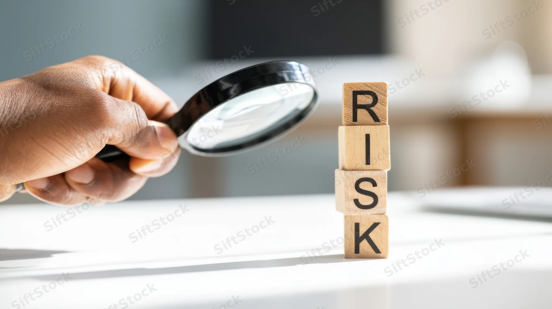 Hand holding magnifying glass over stacked wooden blocks spelling risk on a desk