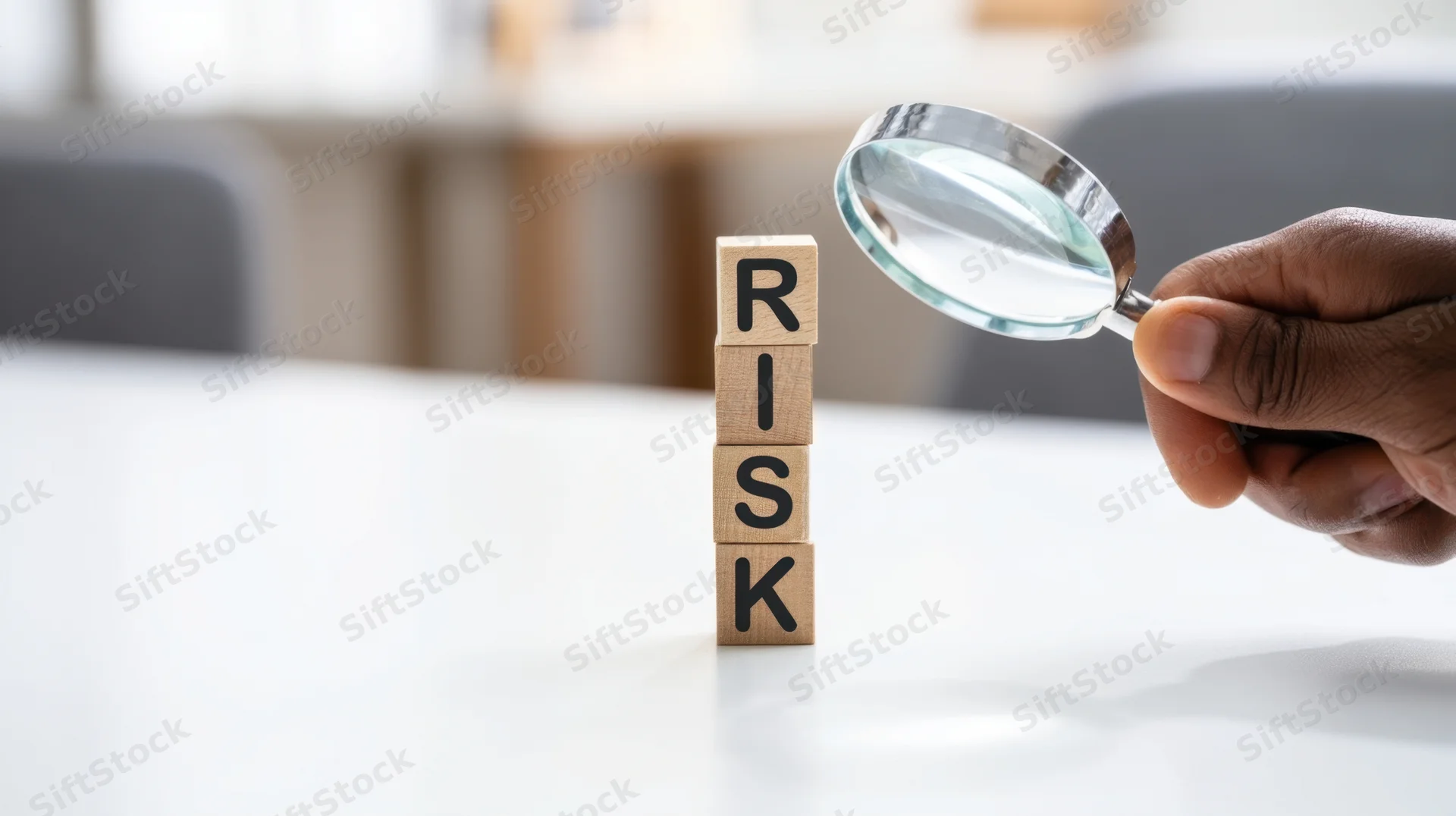 Hand holding magnifying glass over wooden blocks spelling risk on a desk