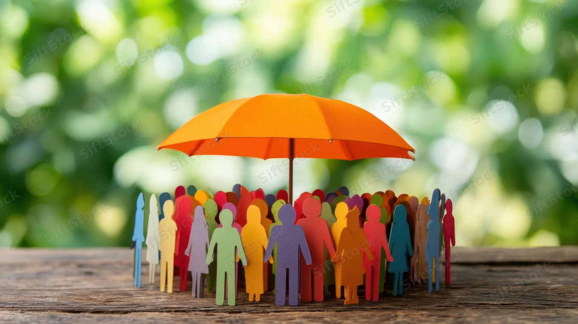 Colorful paper figures gathered under an orange umbrella on a wooden surface with green foliage background