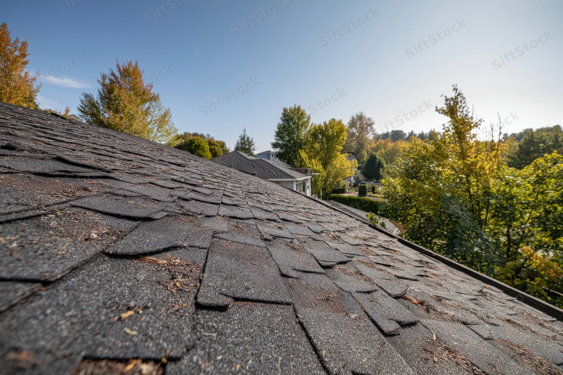 Damaged asphalt shingles on a residential roof in a suburban neighborhood with autumn trees in the background