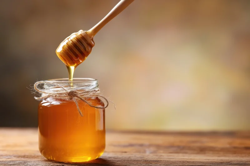 Honey dripping from a wooden dipper into a glass jar on a wooden table