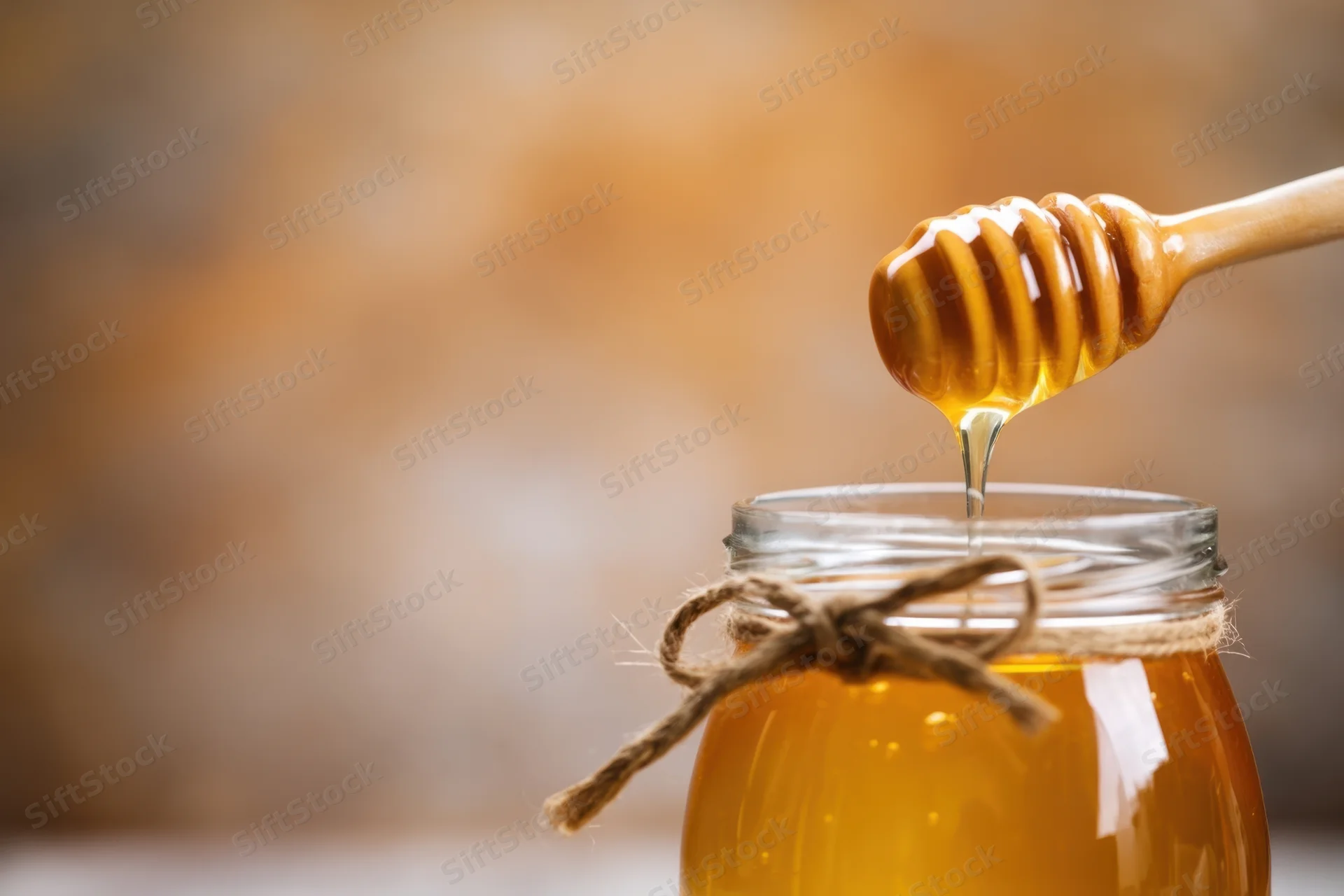 Honey dripping from a wooden dipper into a glass jar tied with twine
