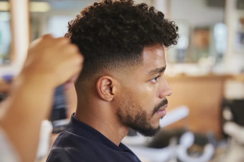 Man receiving haircut in barbershop, side profile view showing curly tapered style