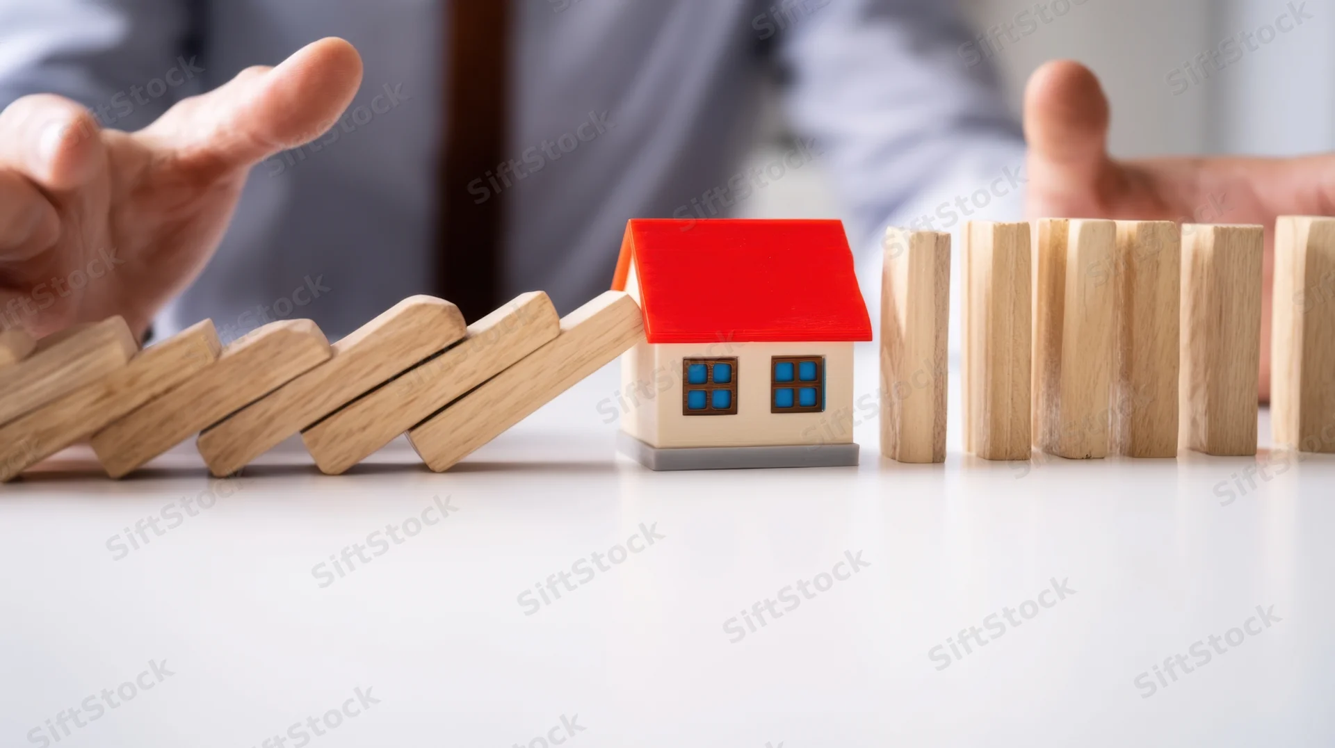 Hands stopping falling dominoes to protect a miniature house on a desk
