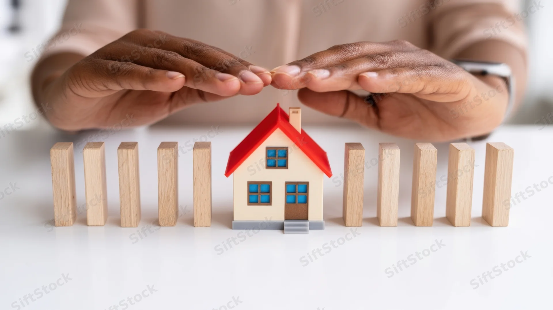 Hands shielding a miniature house from falling wooden blocks on a white surface