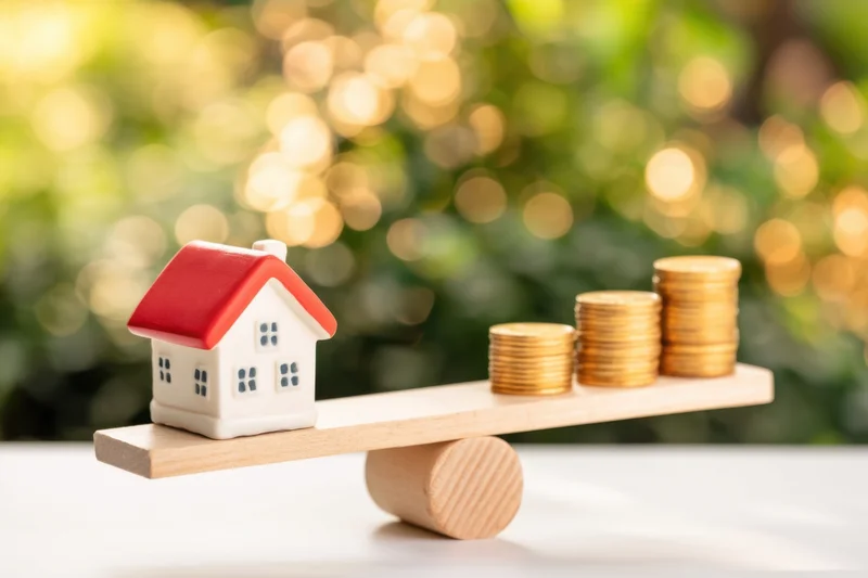 Miniature house and stacks of coins balanced on a wooden seesaw against a blurred green background