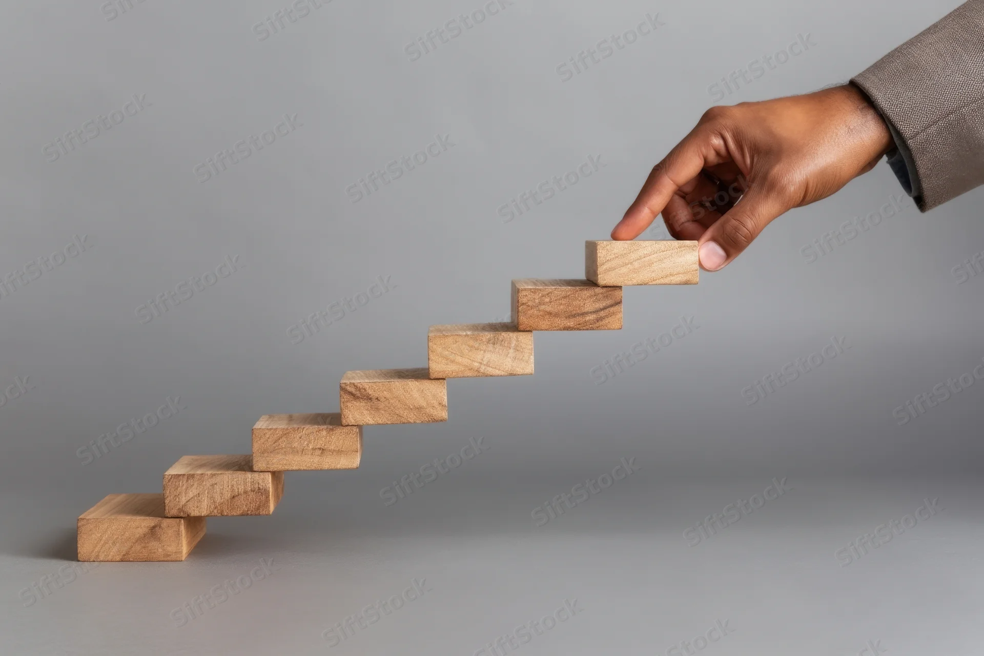 Hand placing wooden block on ascending staircase against gray background