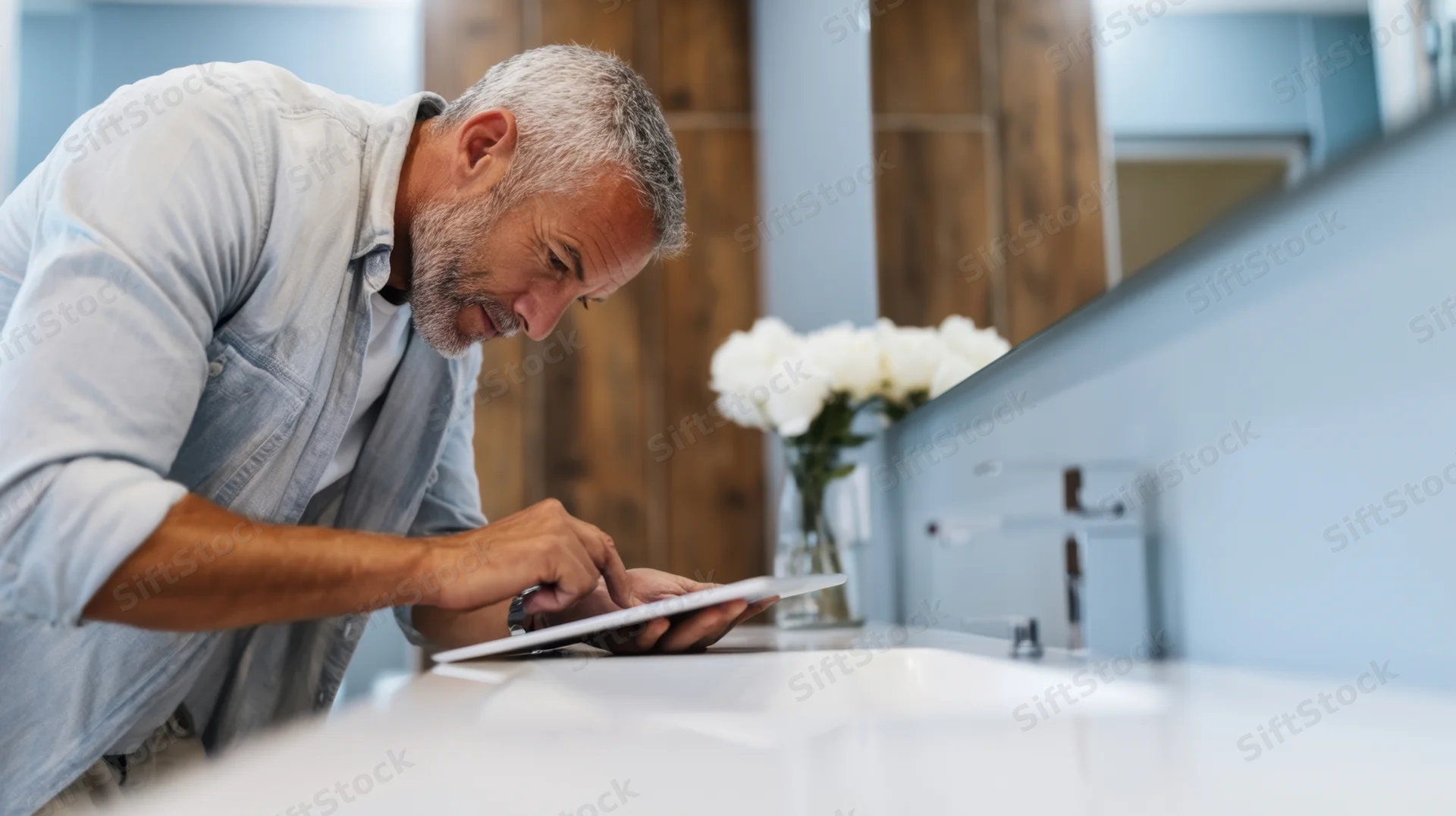 Senior man using tablet at bathroom countertop near sink and mirror