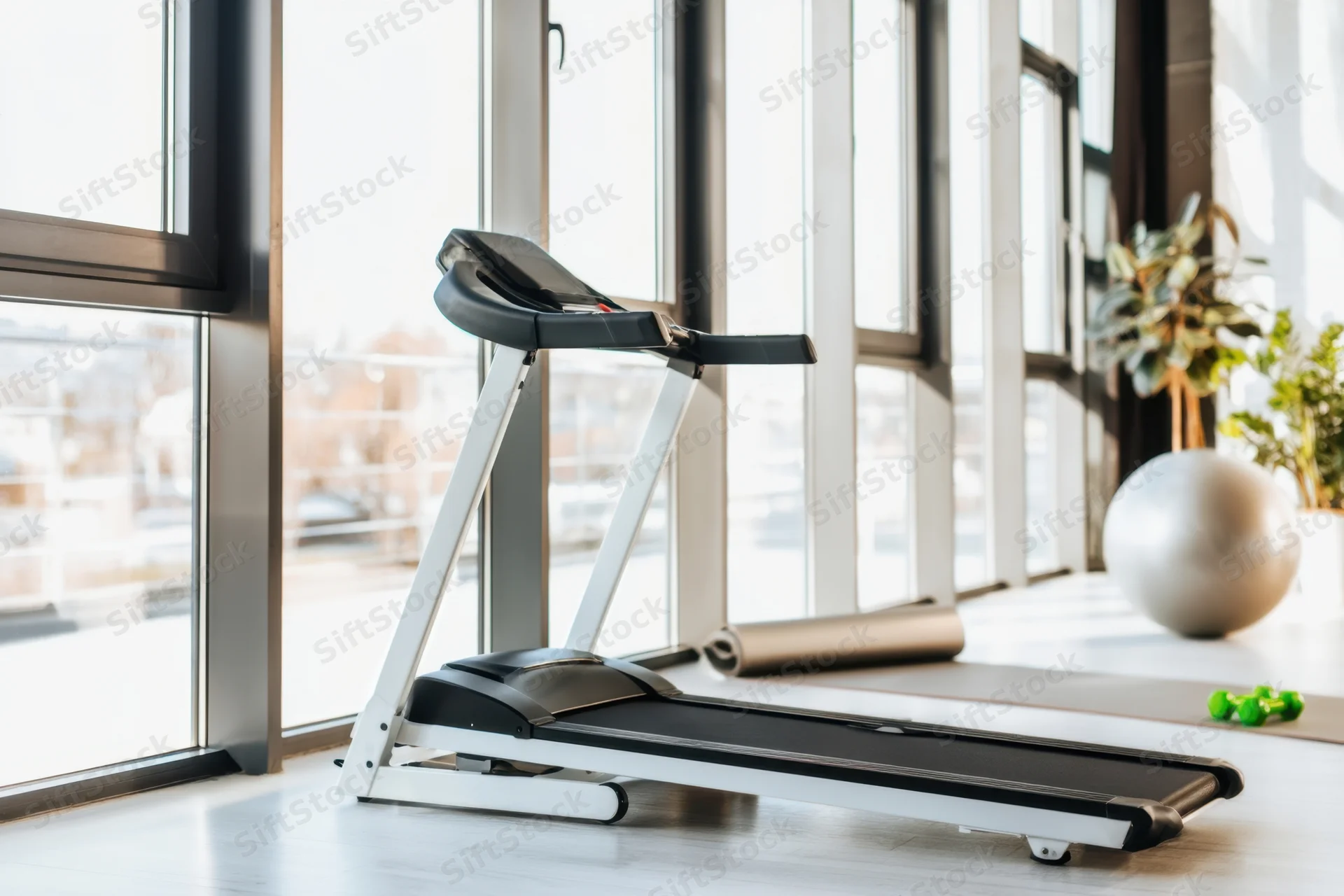 Modern treadmill in a bright home gym space with large windows and fitness accessories nearby