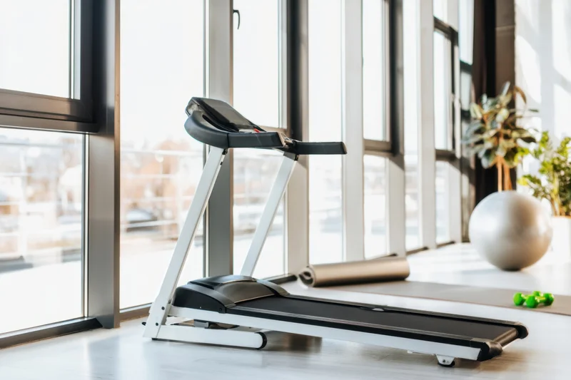 Modern treadmill in a bright home gym space with large windows and fitness accessories nearby