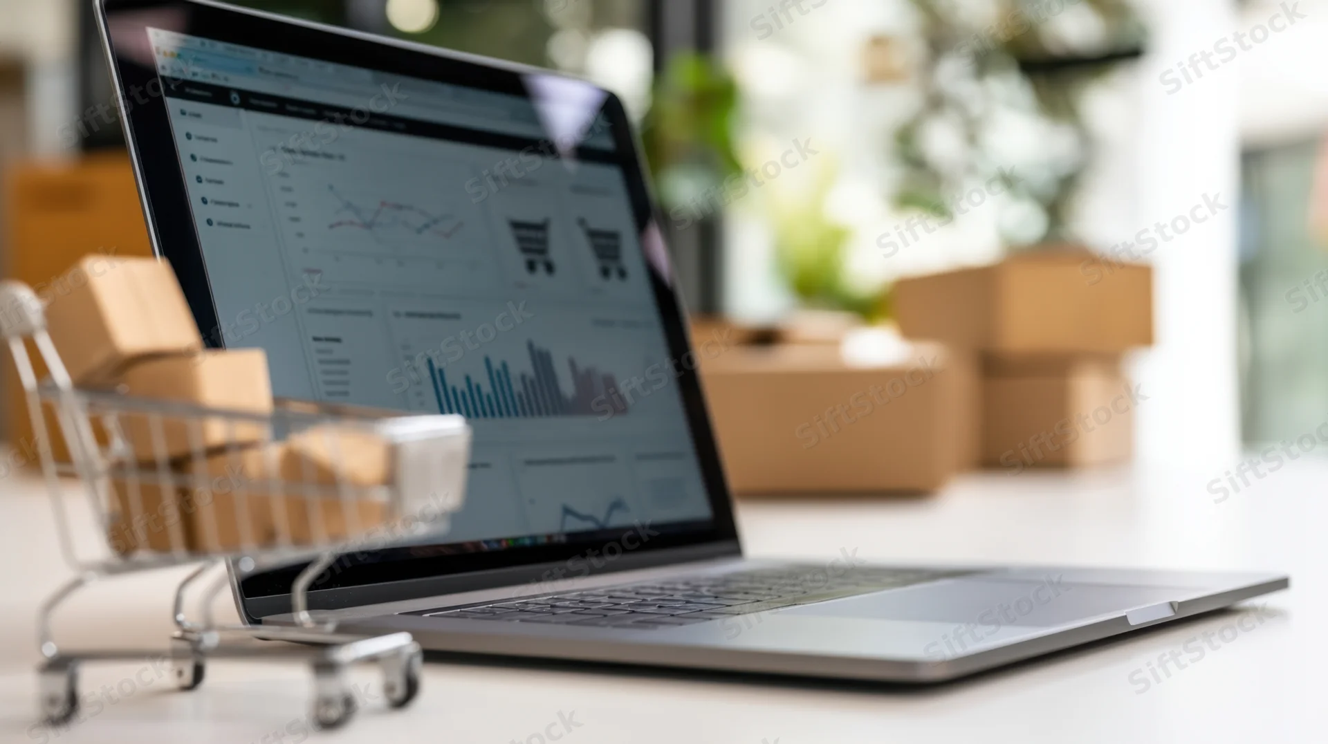 Laptop displaying e-commerce analytics dashboard with miniature shopping cart and cardboard boxes on desk