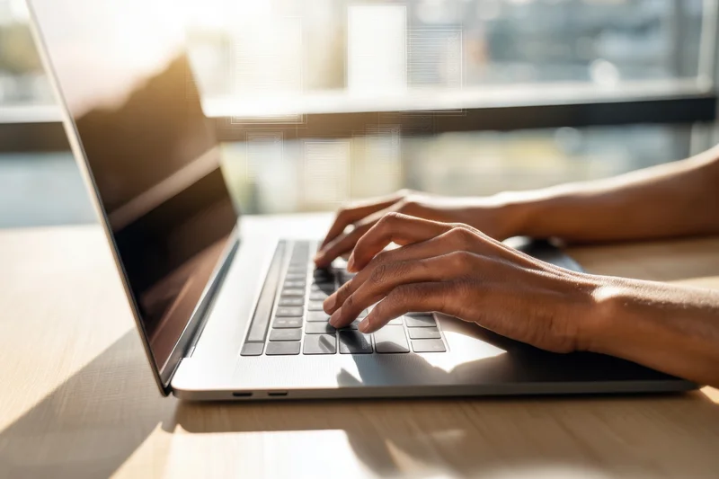 Hands typing on a laptop at a desk in a bright office space