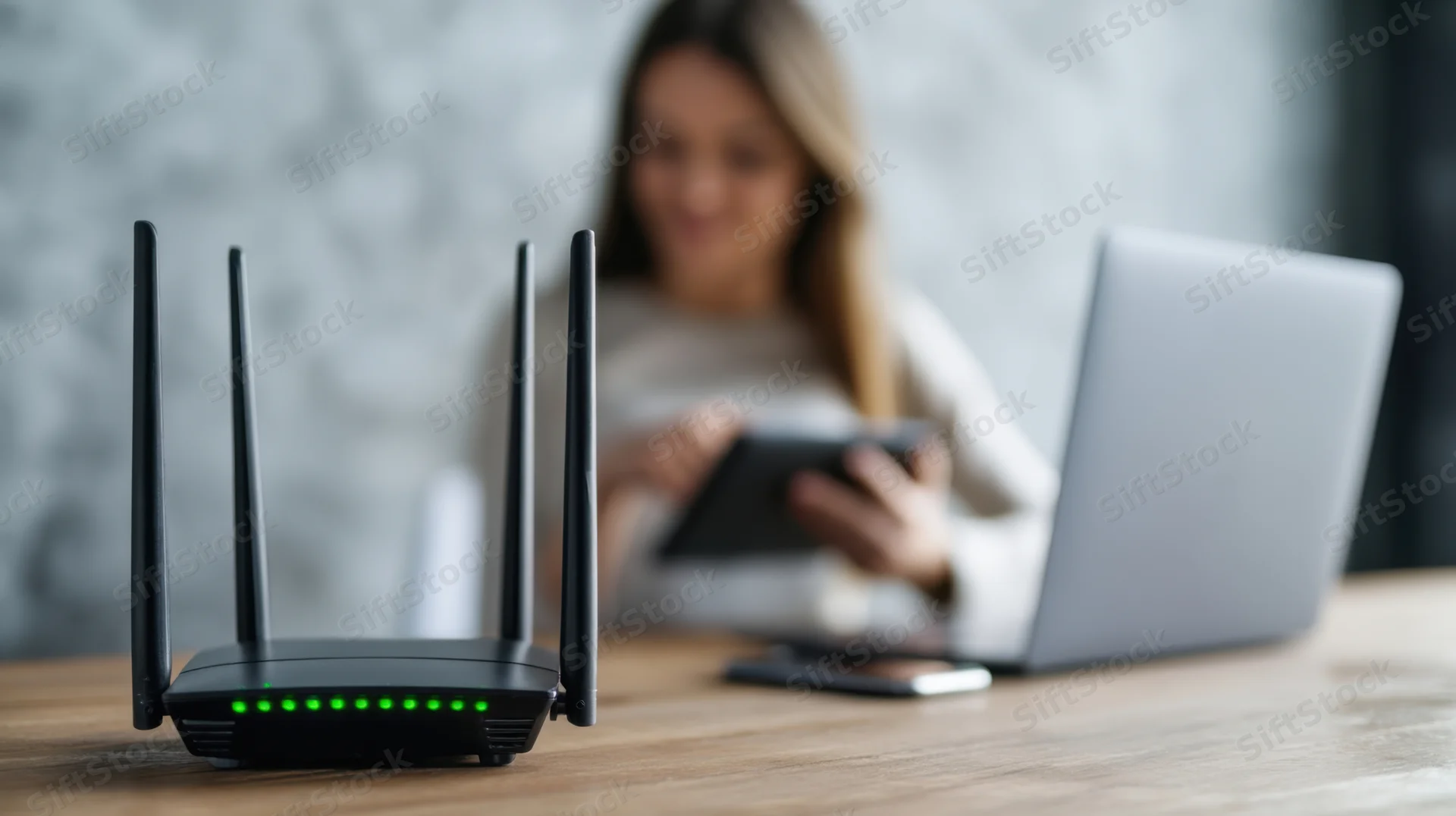Black wi-fi router with green led lights on wooden table, woman using tablet in background, laptop nearby