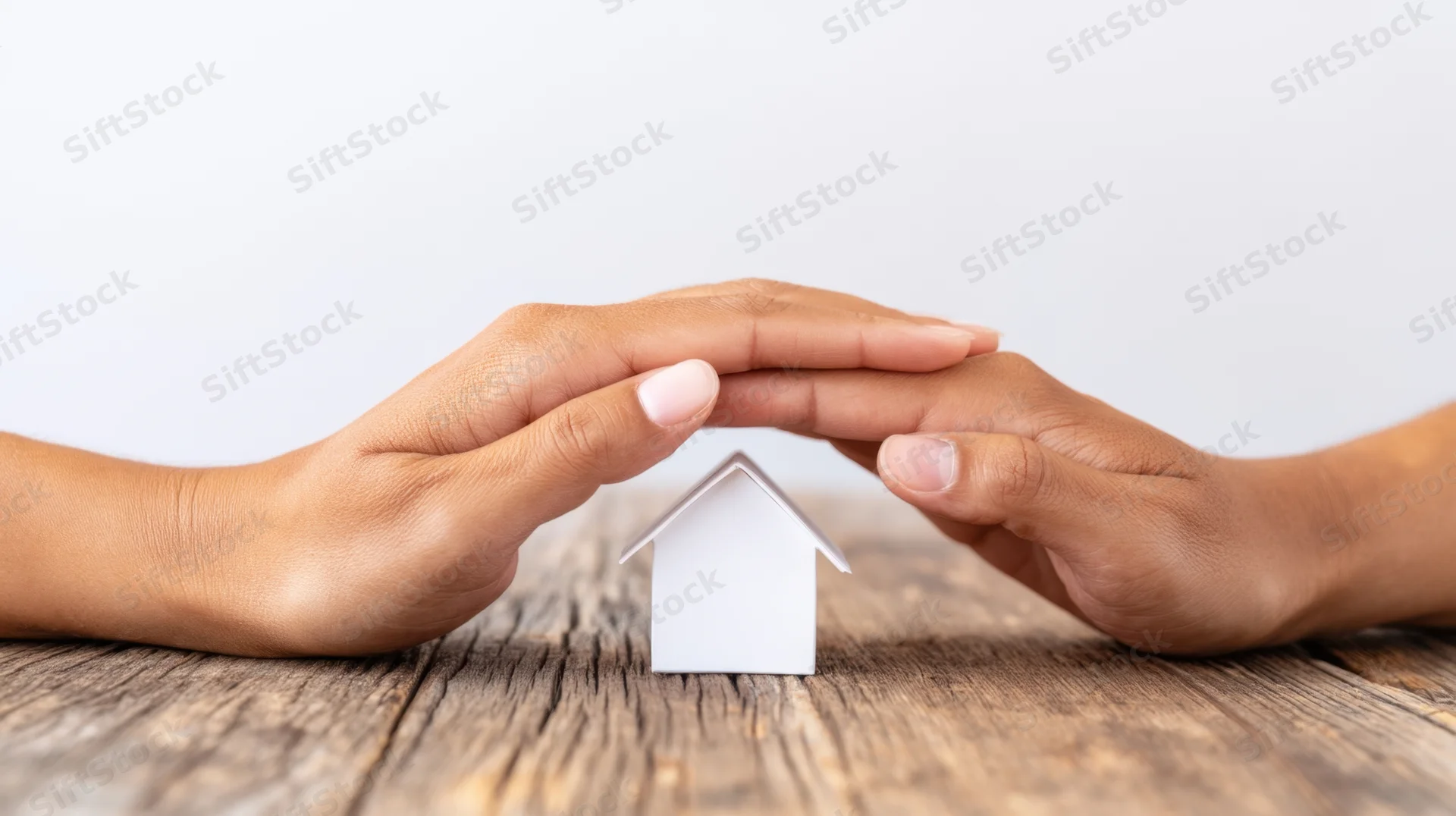 Hands forming a protective arch over a small white house model on a wooden table