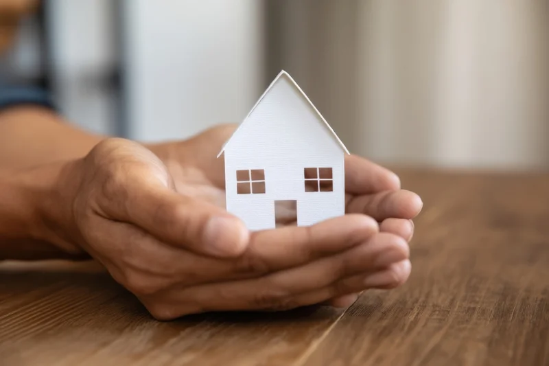 Hands holding a white paper house model on a wooden table