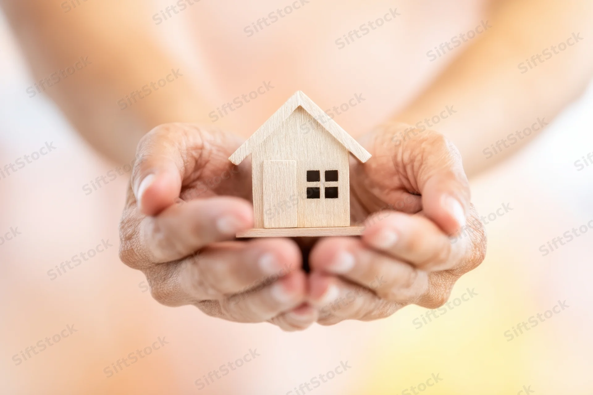 Hands cradling a small wooden house model against a soft blurred background