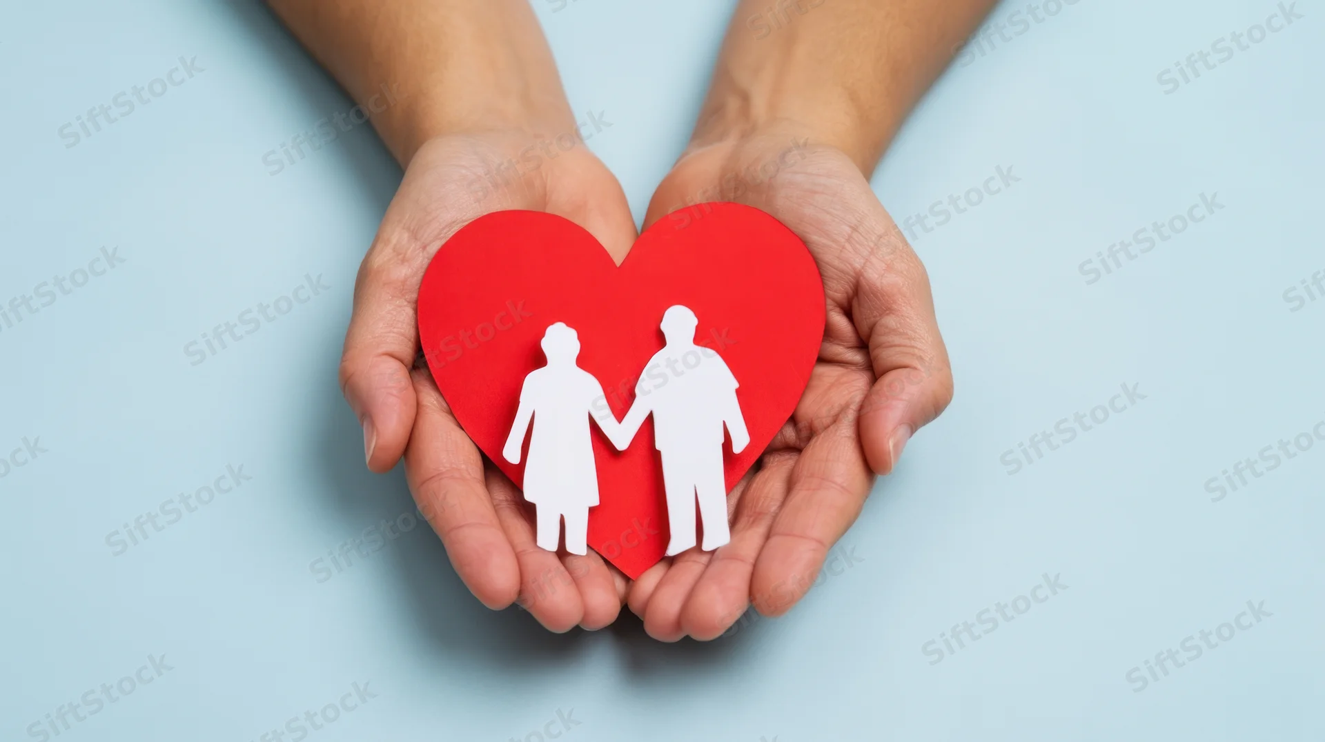 Hands holding a red paper heart with white cutout figures of a couple on a light blue background