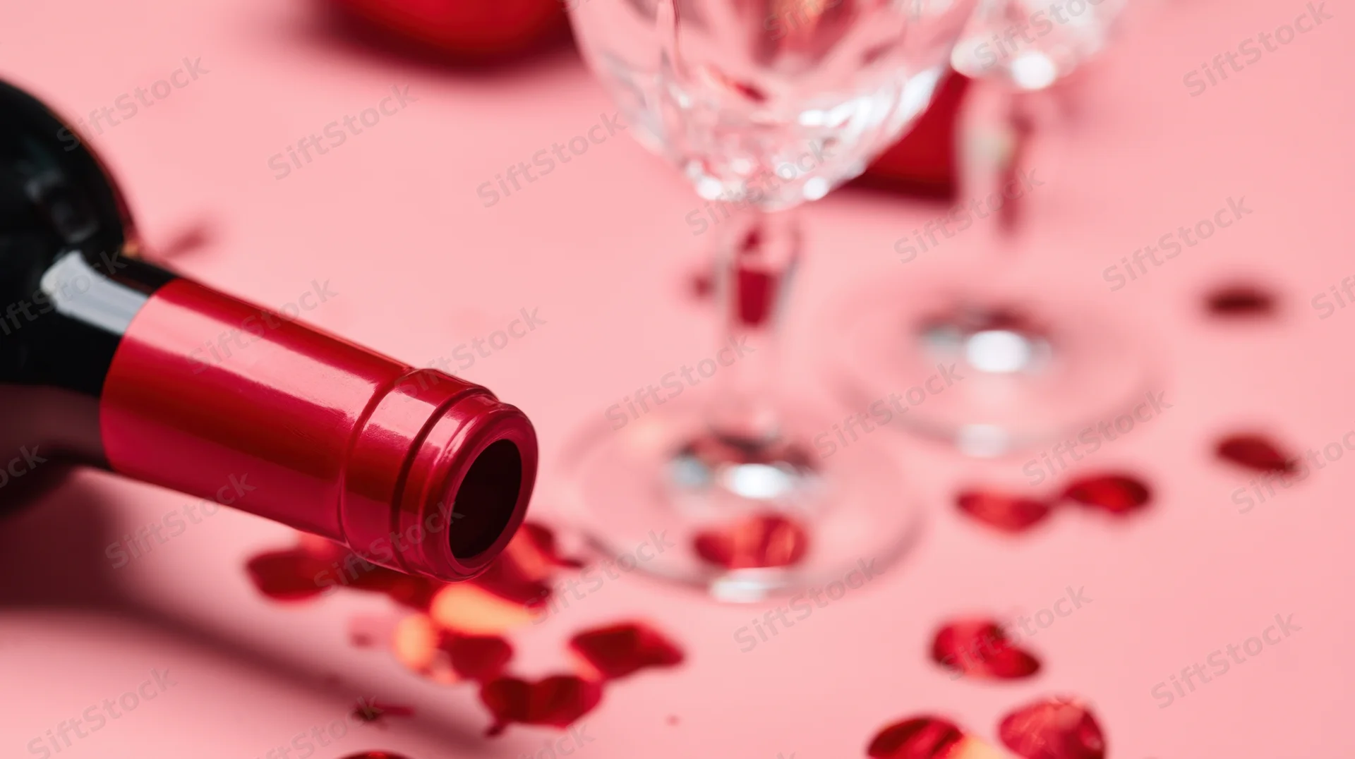 Red wine bottle lying on pink surface with rose petals and wine glasses nearby