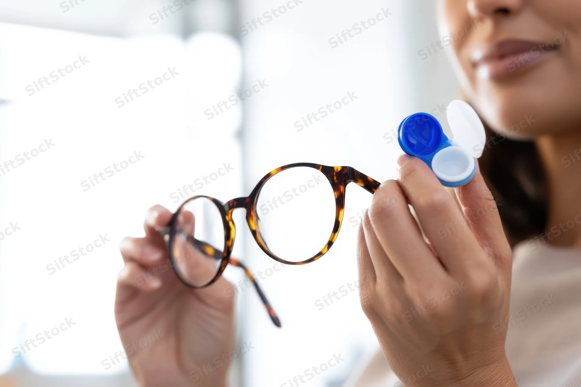 Woman holding tortoiseshell eyeglasses and open contact lens case indoors