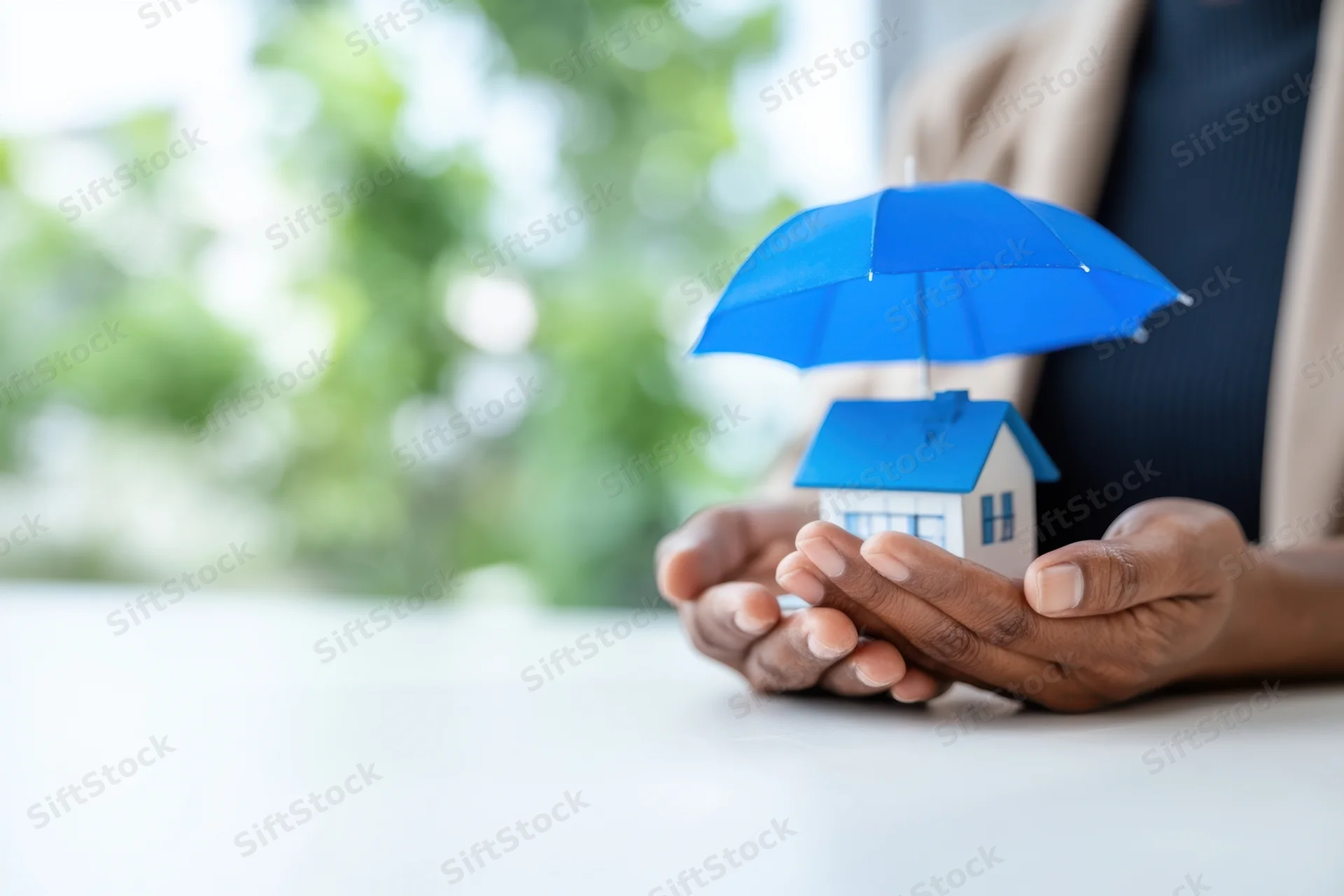 Hands holding a miniature house under a blue umbrella in an office setting