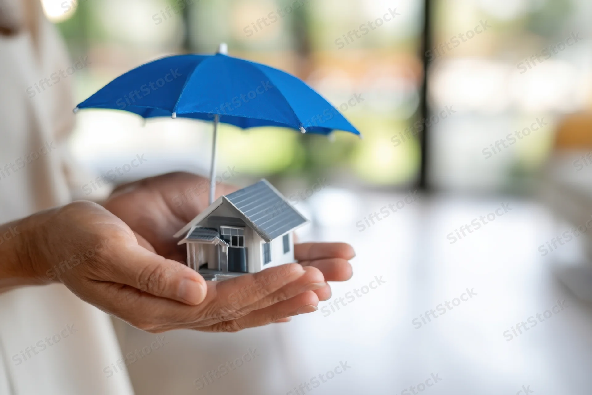 Hands holding a miniature house under a blue umbrella indoors