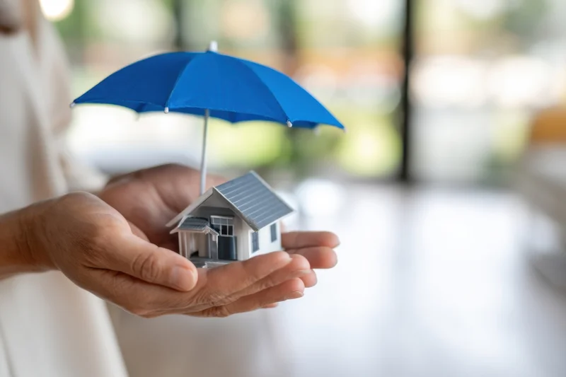 Hands holding a miniature house under a blue umbrella indoors