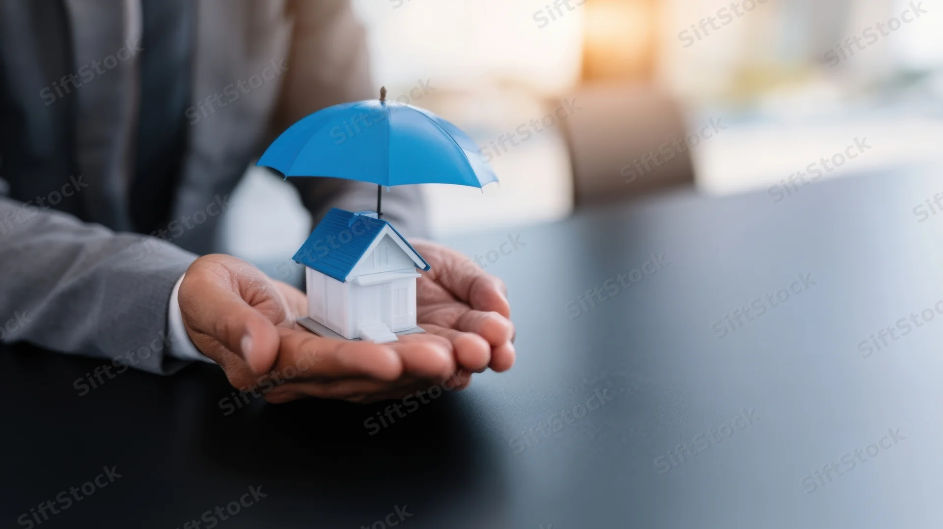 Businessman holding a miniature house under a blue umbrella on a desk