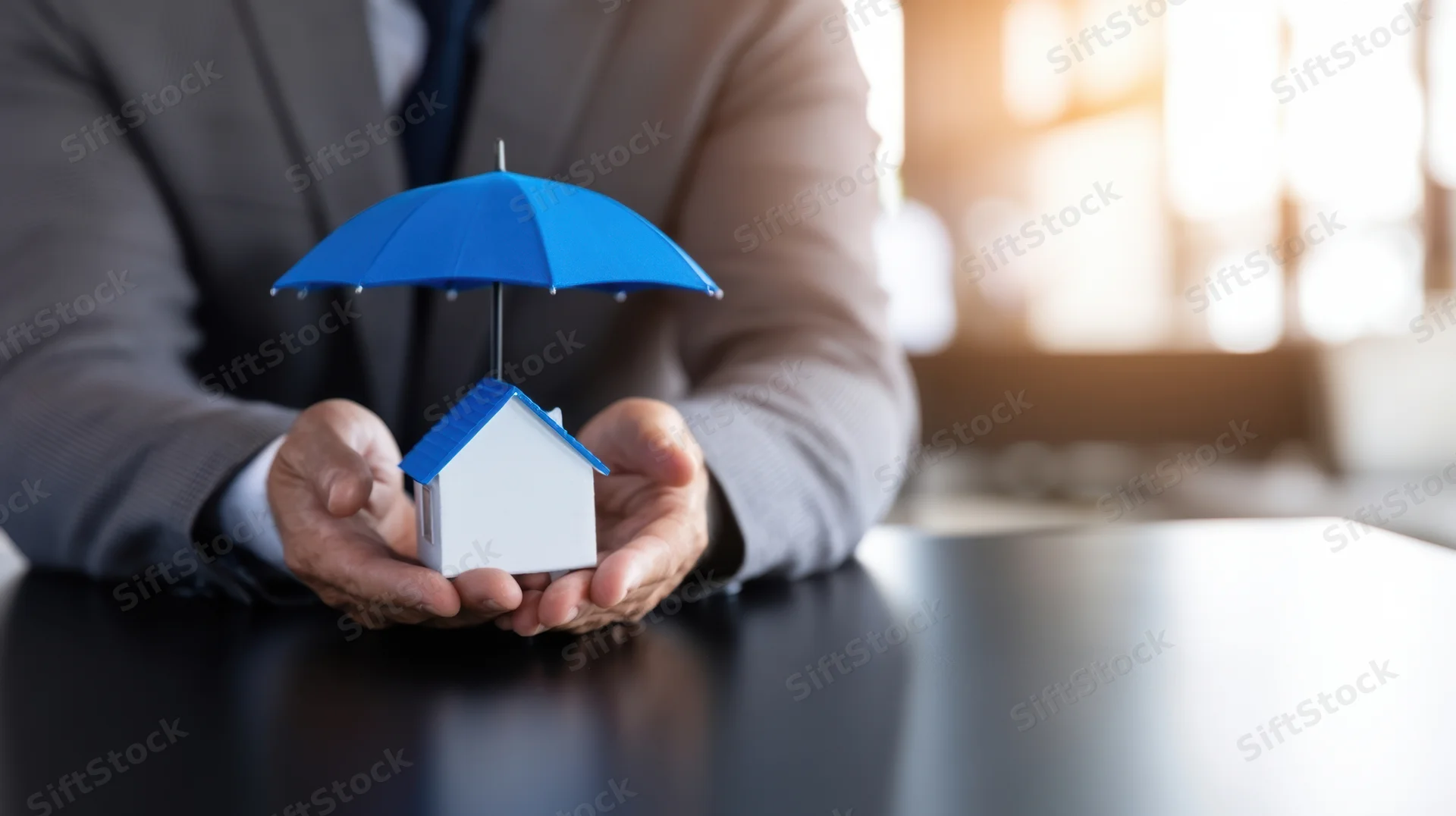 Man in suit holding miniature house under blue umbrella on office desk